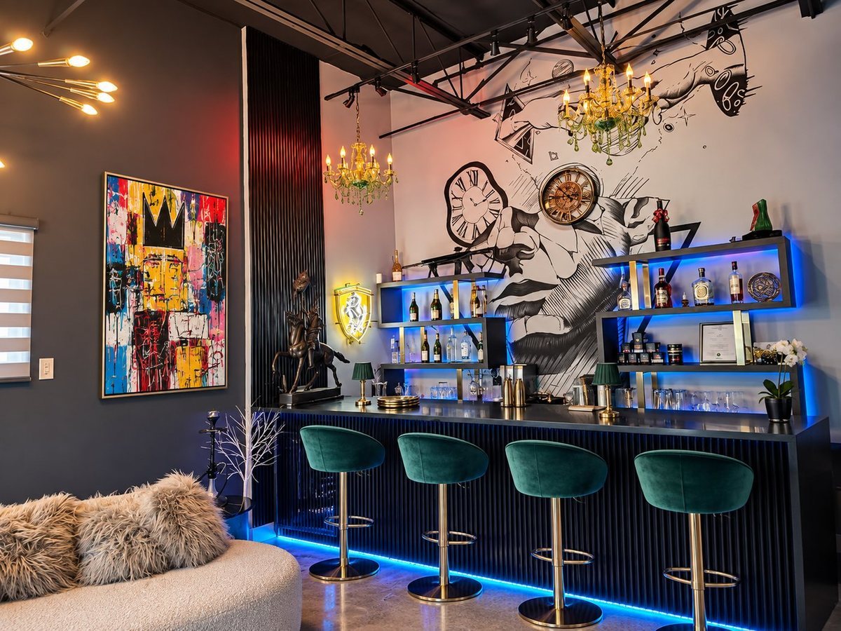 A bar with four green velvet stools, lit bottle shelves, chandeliers and a wall mural