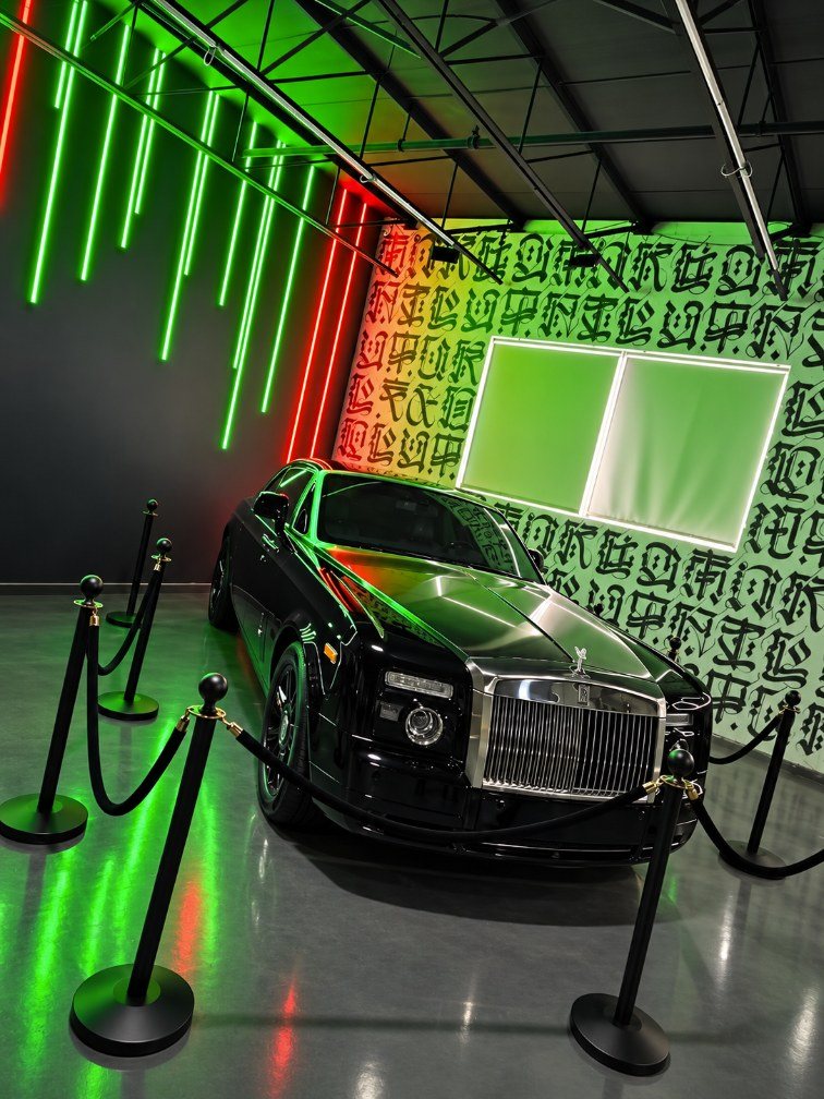 A black Rolls-Royce Phantom Coupe behind stanchions under green and red neon light